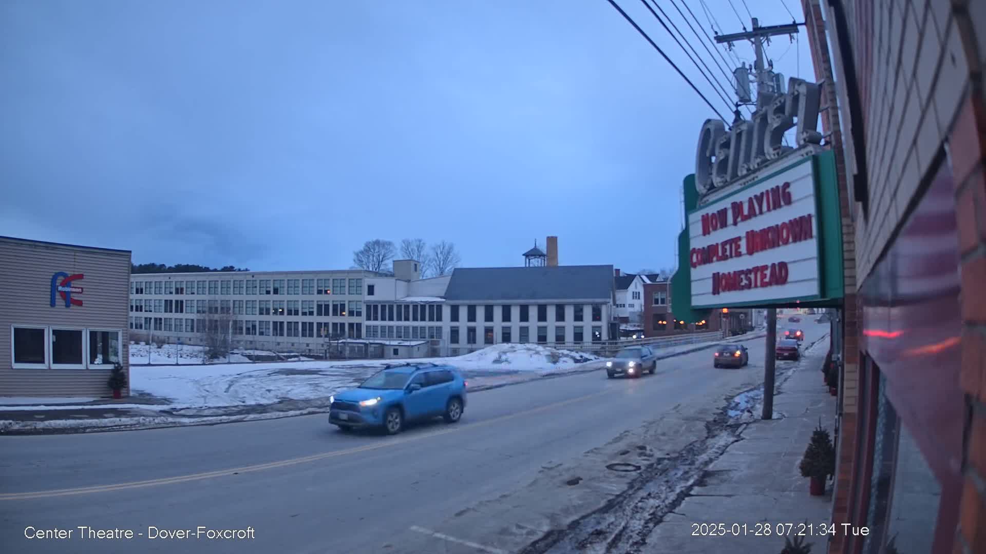 A street scene on a twilight evening shows a few cars driving past buildings, including a multi-story structure and a smaller one with a marquee.