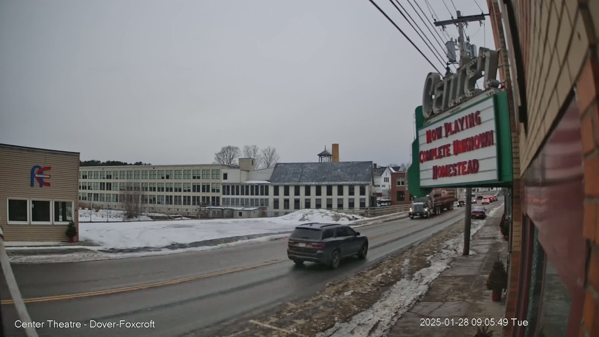 A street scene on an overcast day shows a road with cars driving past several buildings, including a large factory and a smaller building with a movie marquee.