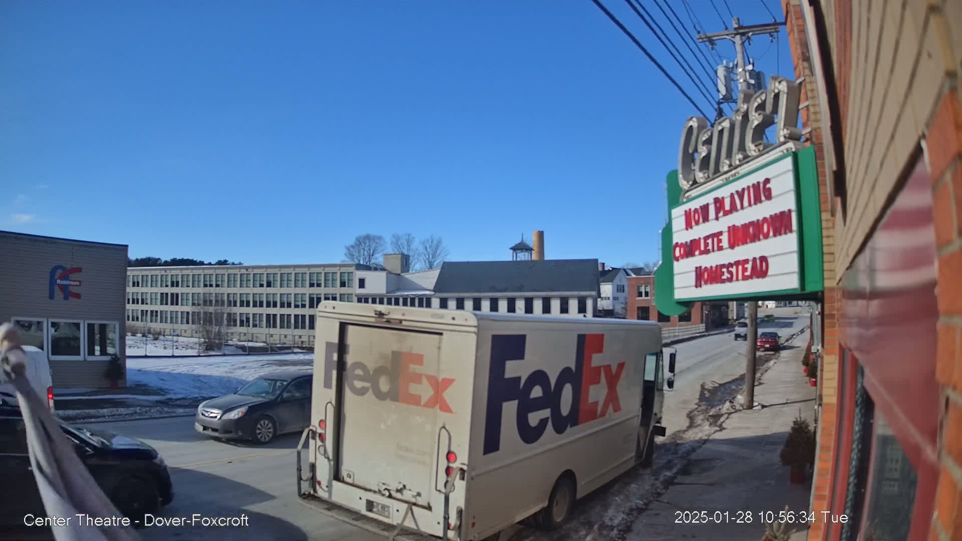 A FedEx truck is parked on a street on a sunny day, with cars driving past and buildings in the background, including a multi-story building and a movie theater marquee.