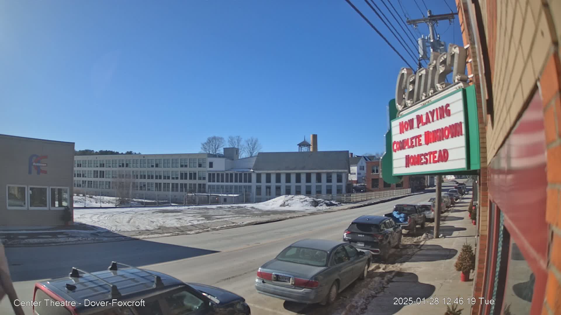 A sunny day reveals a street scene with parked cars, a large building complex, and a movie theater marquee.