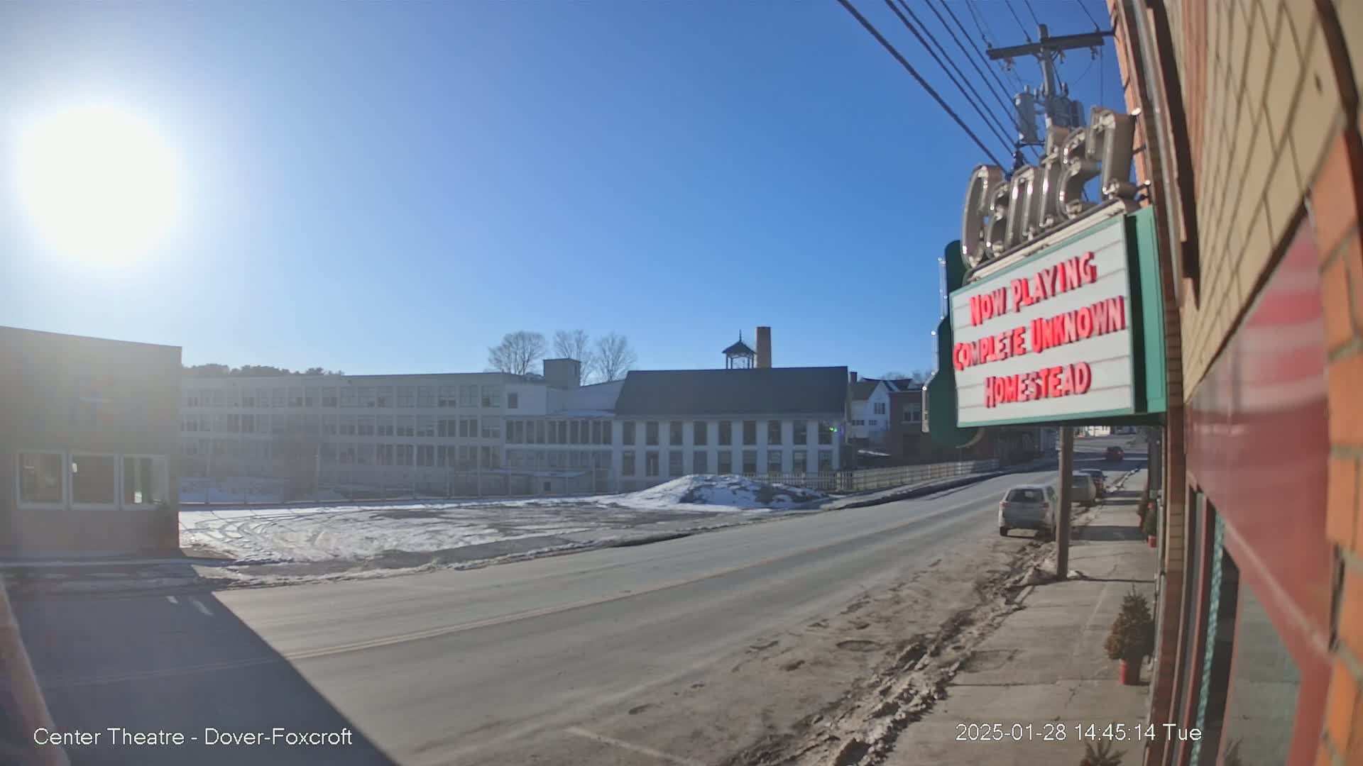 A sunny day reveals a street scene with a large, multi-story building across the street, partially snow-covered ground, and a marquee displaying movie titles.