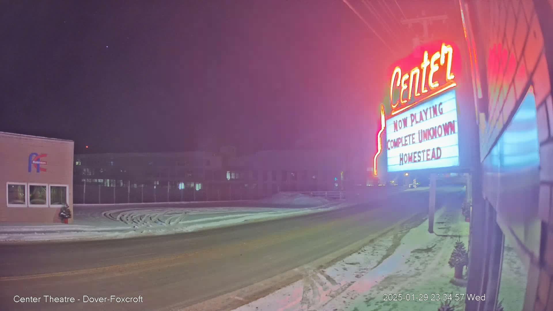 A snow-covered road at night leads to a movie theater marquee advertising "Complete Unknown" and "Homestead."