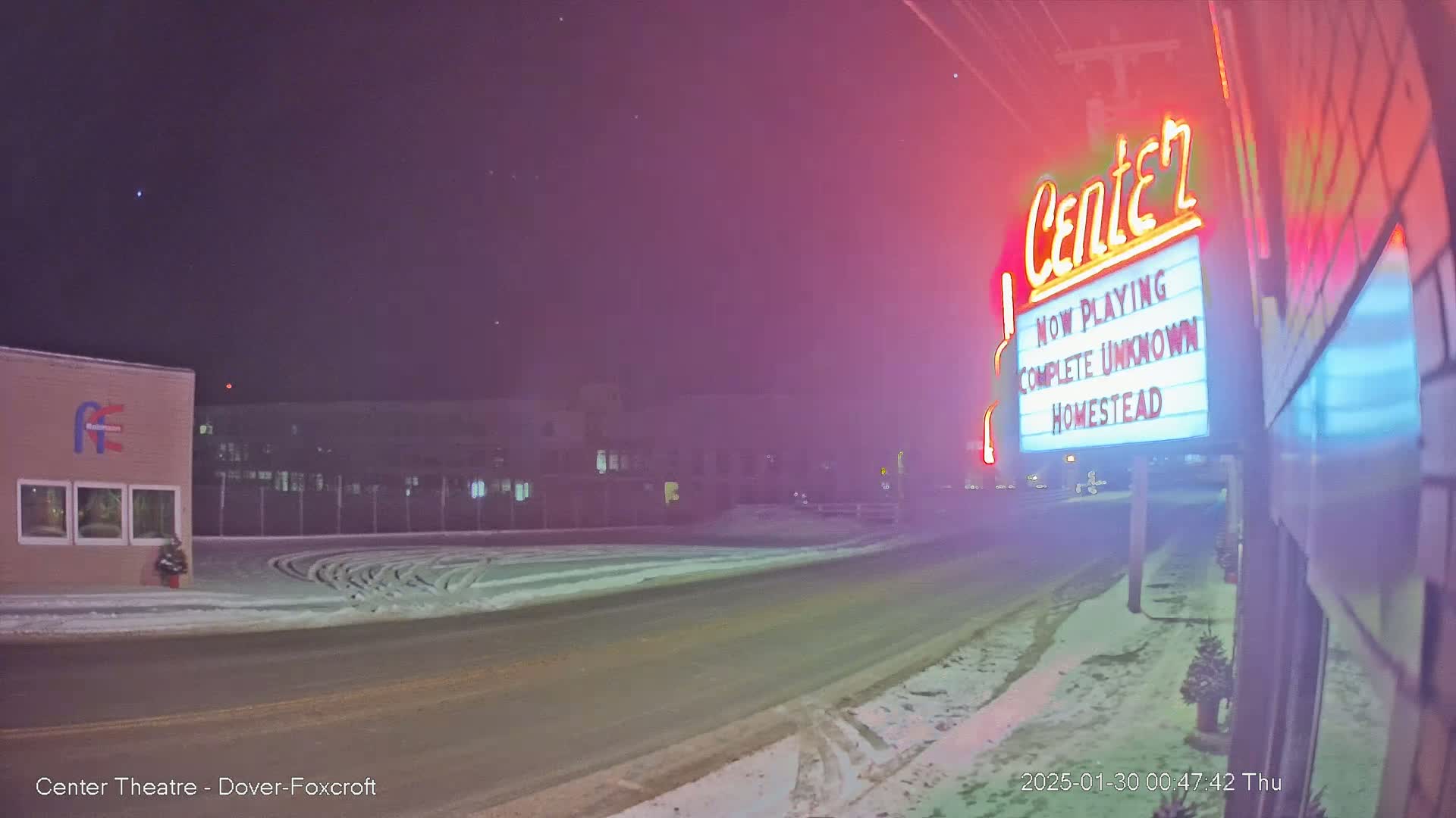 A snow-covered road at night leads to a movie theater marquee displaying "Complete Unknown" and "Homestead."