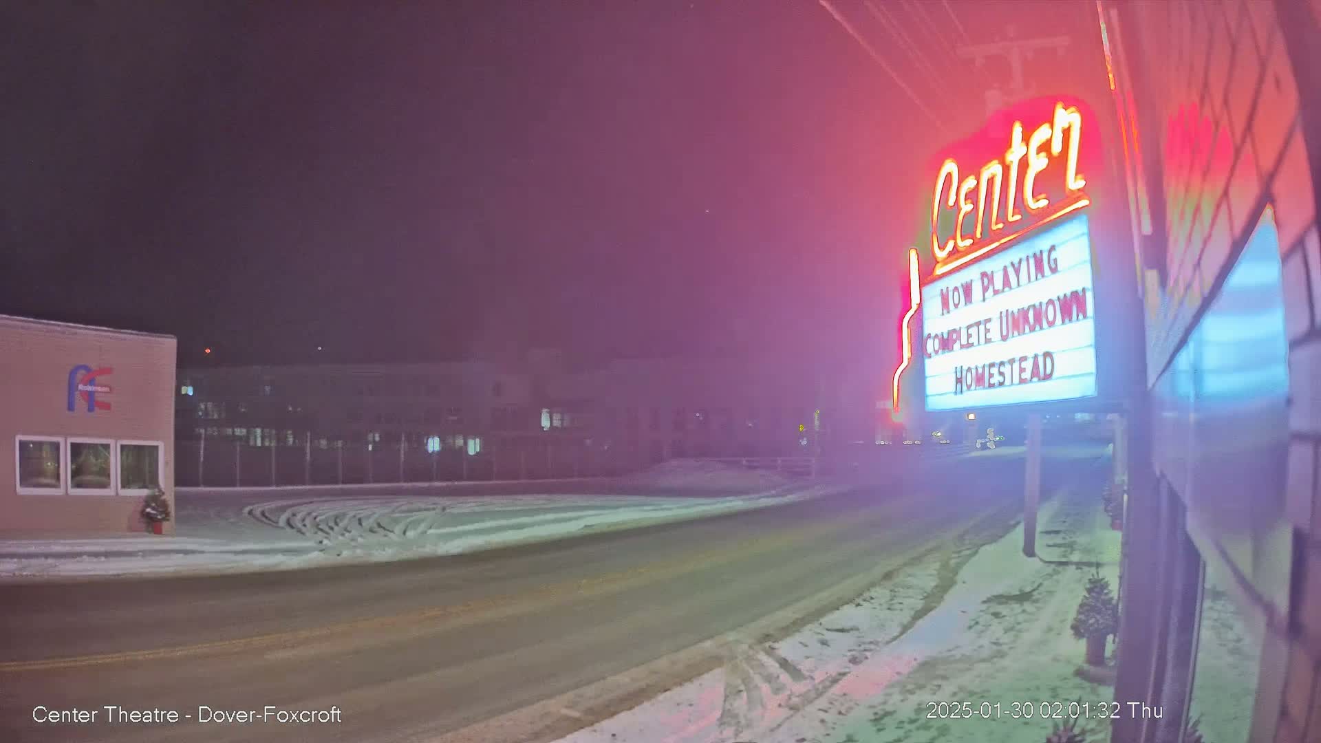 A snow-covered street at night leads to a movie theater marquee advertising "Complete Unknown" and "Homestead," under a hazy, dimly lit sky.