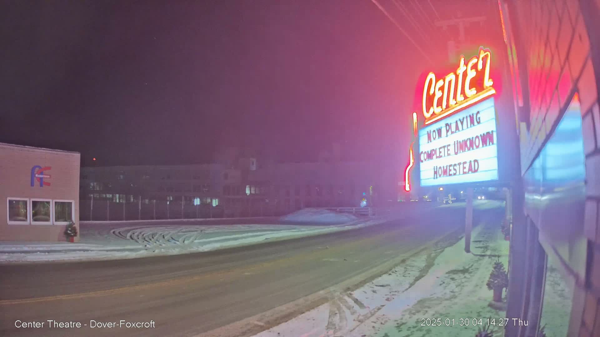 A snow-covered street at night is seen with a movie theater marquee displaying "Complete Unknown" and "Homestead," lit up against a hazy, dark sky.