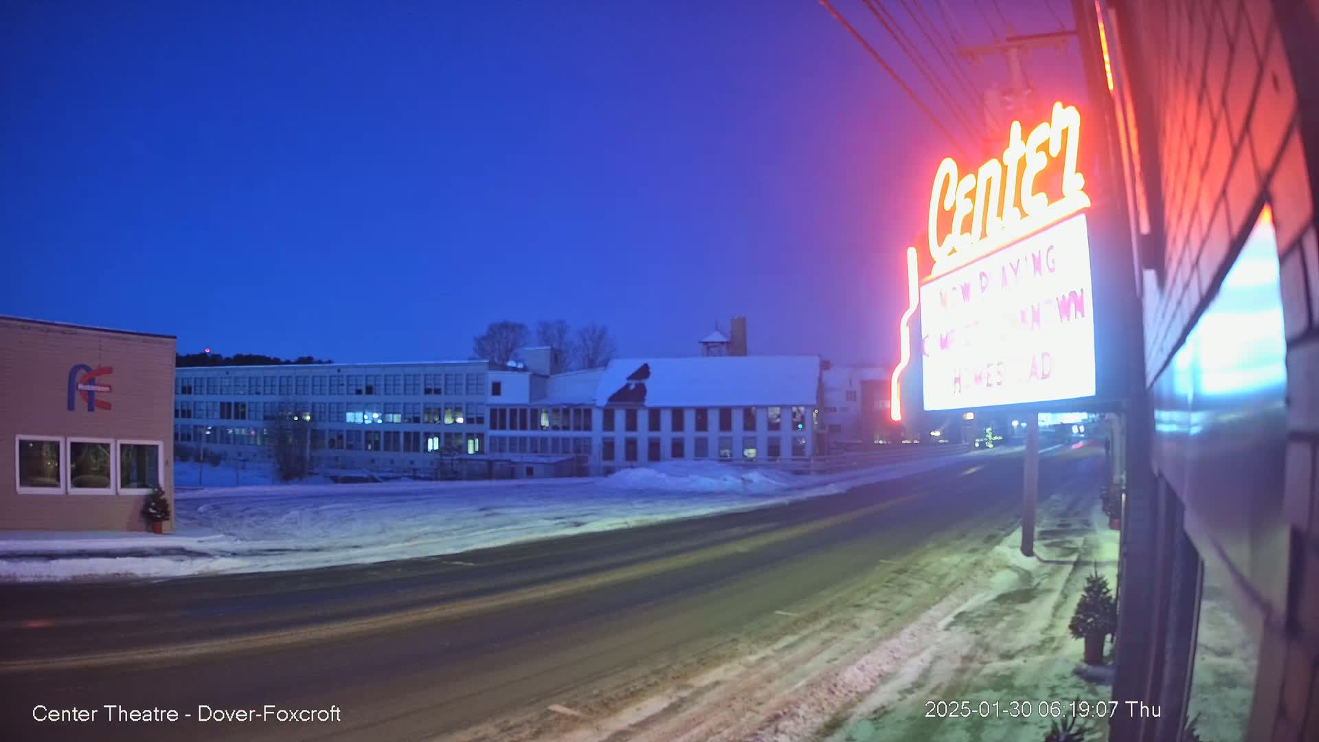 A snow-covered street at twilight is shown with a neon sign for a theater and several buildings in the background.