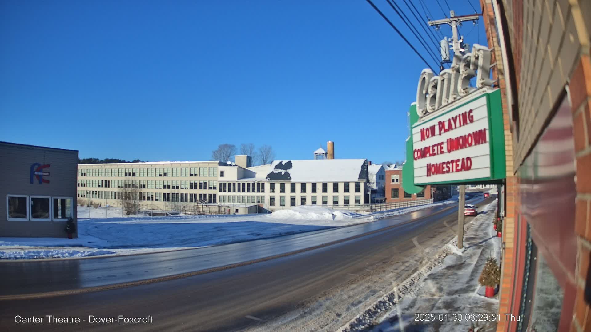 A snow-covered street scene on a sunny day shows a large factory building and a movie theater marquee.