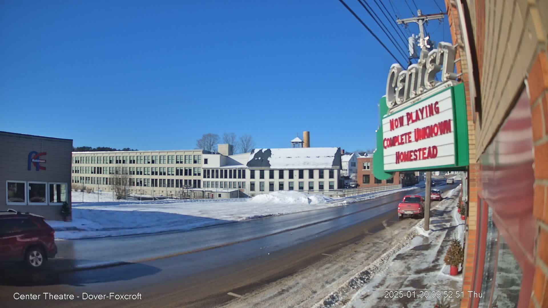 A snow-covered street scene on a sunny day shows several buildings, including a large factory and a theater marquee, with a few cars driving along the road.