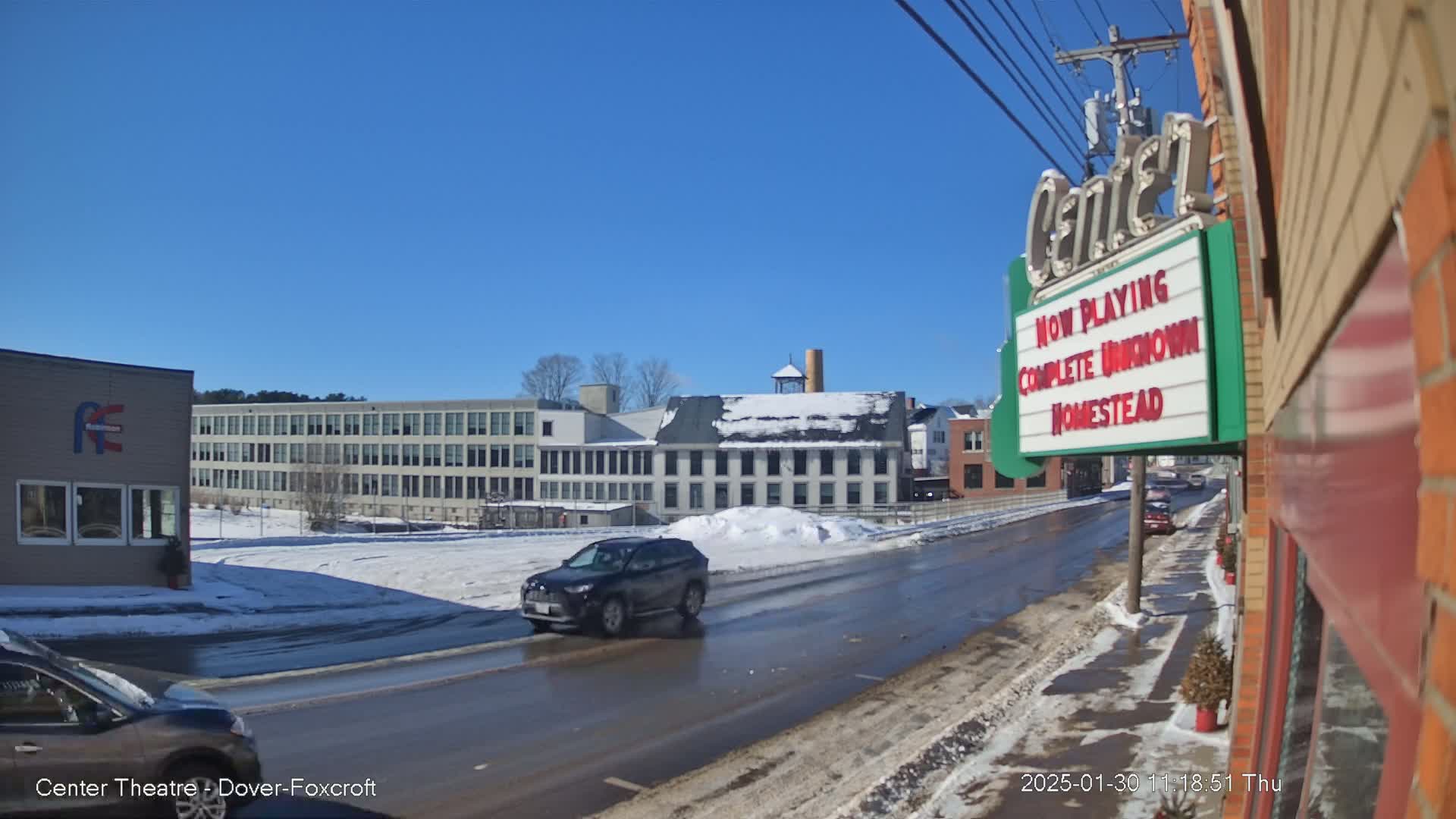 A sunny winter day shows a street scene with melting snow and ice on the road, passing cars, and a multi-story building in the background.