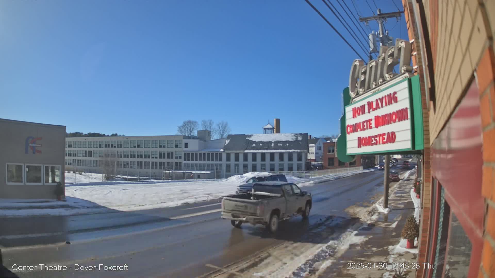 A snow-covered street scene on a sunny day shows a pickup truck driving past buildings, including a large school and a factory.