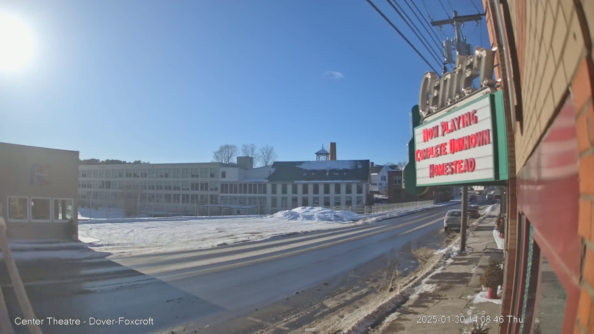 A sunny winter day reveals a street scene with snow-covered ground, buildings including a school and a multi-story factory, and a movie theater marquee.