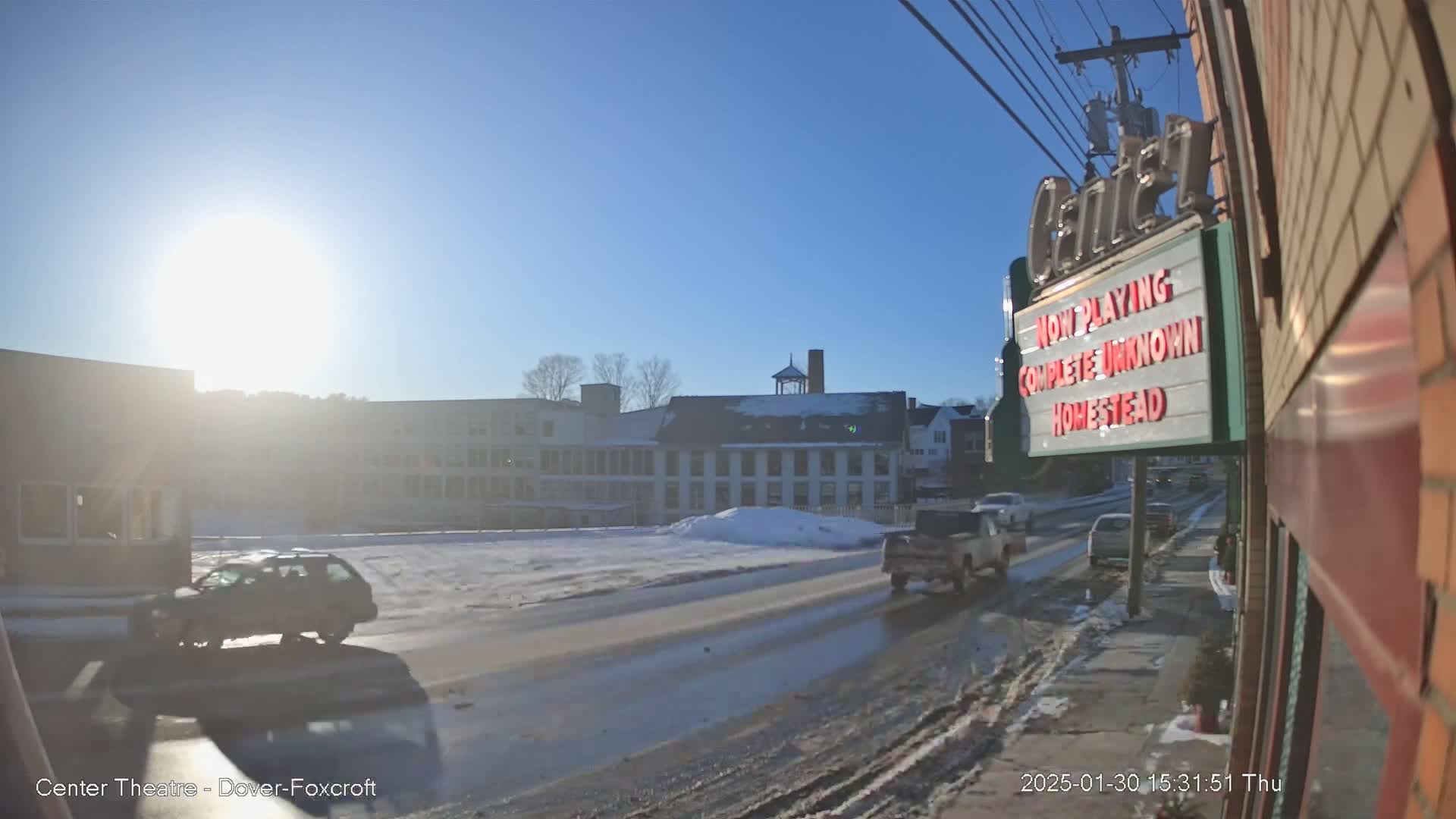 A sunny, snowy day scene shows a street with cars and buildings, including a multi-story building and a theater marquee.