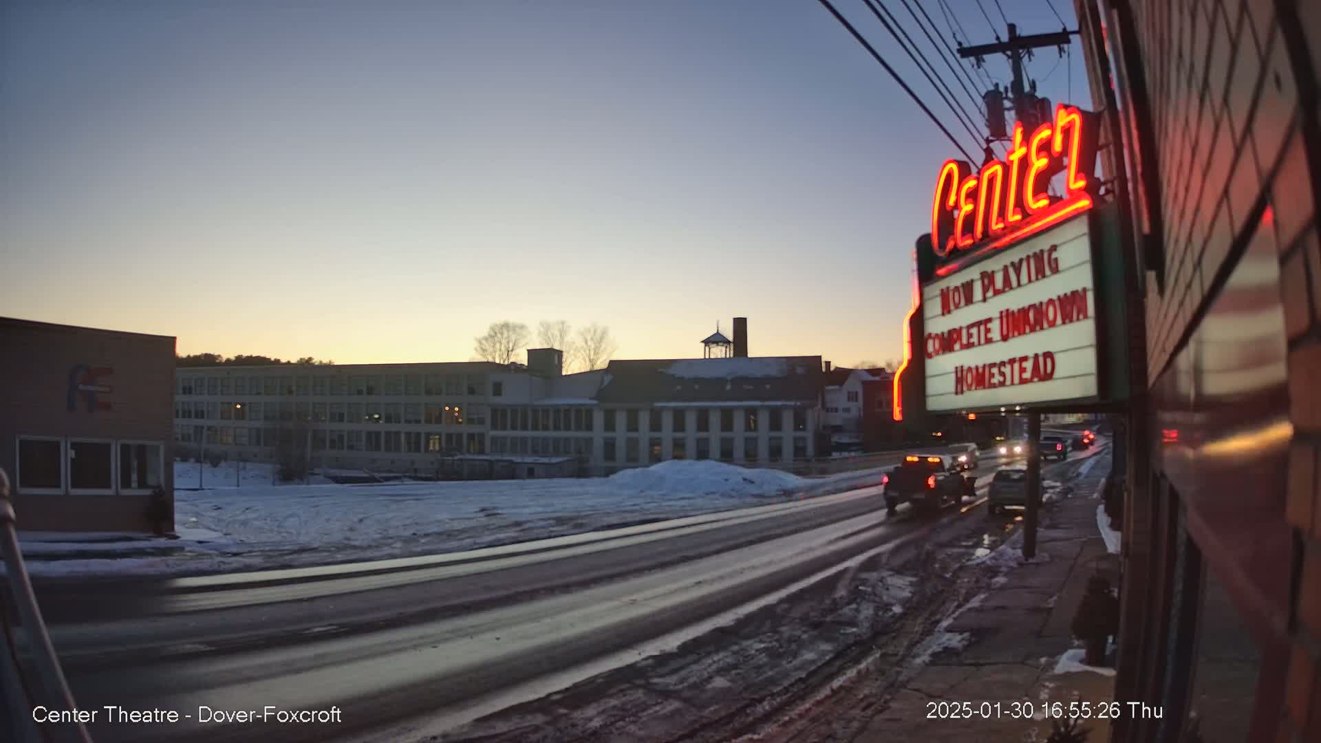 A snow-covered street at dusk shows a large building across the street from a movie theater marquee displaying the films "Complete Unknown" and "Homestead."