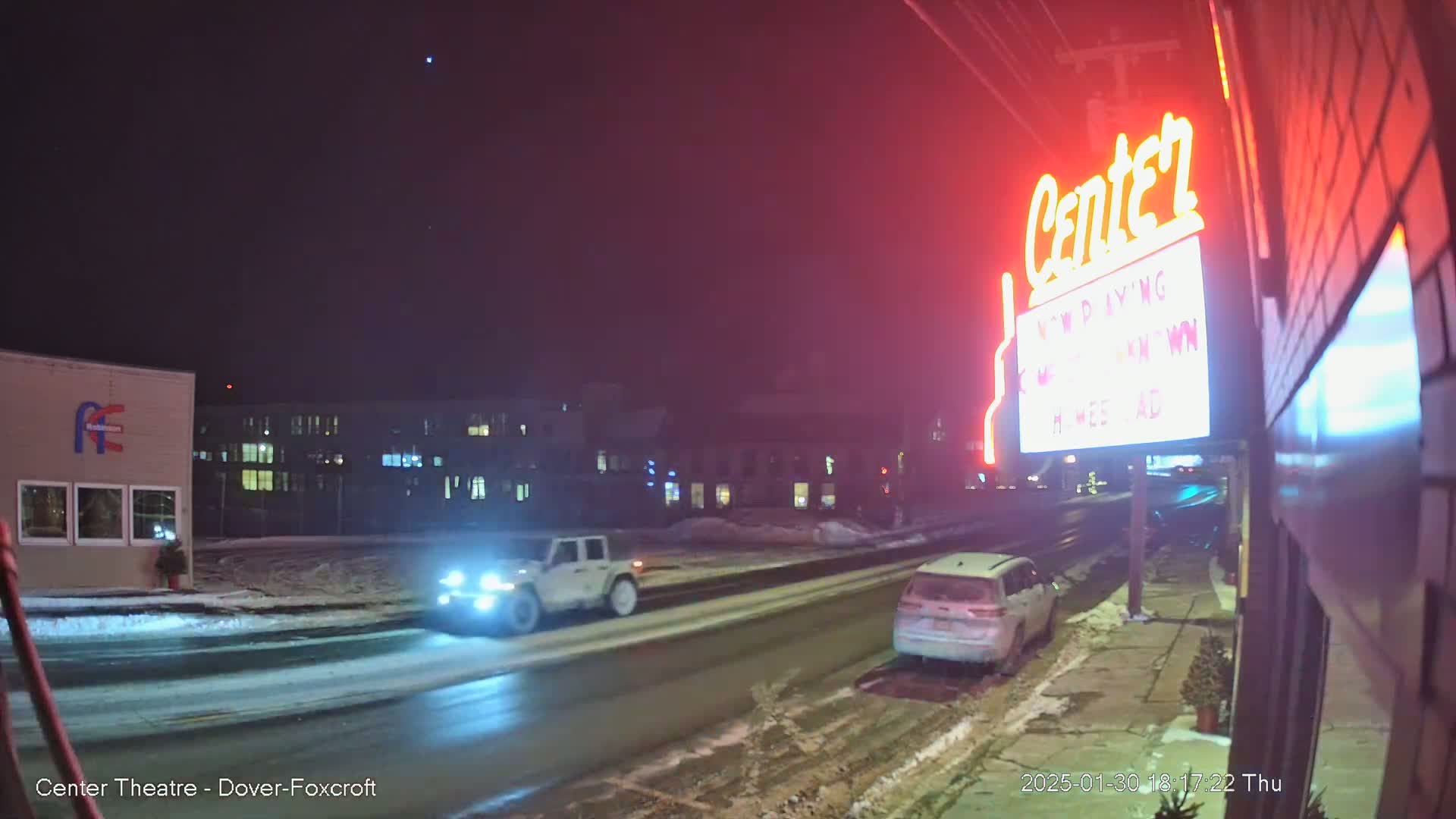 A nighttime street scene shows a snow-covered road with two vehicles passing by a movie theater with a lit marquee.