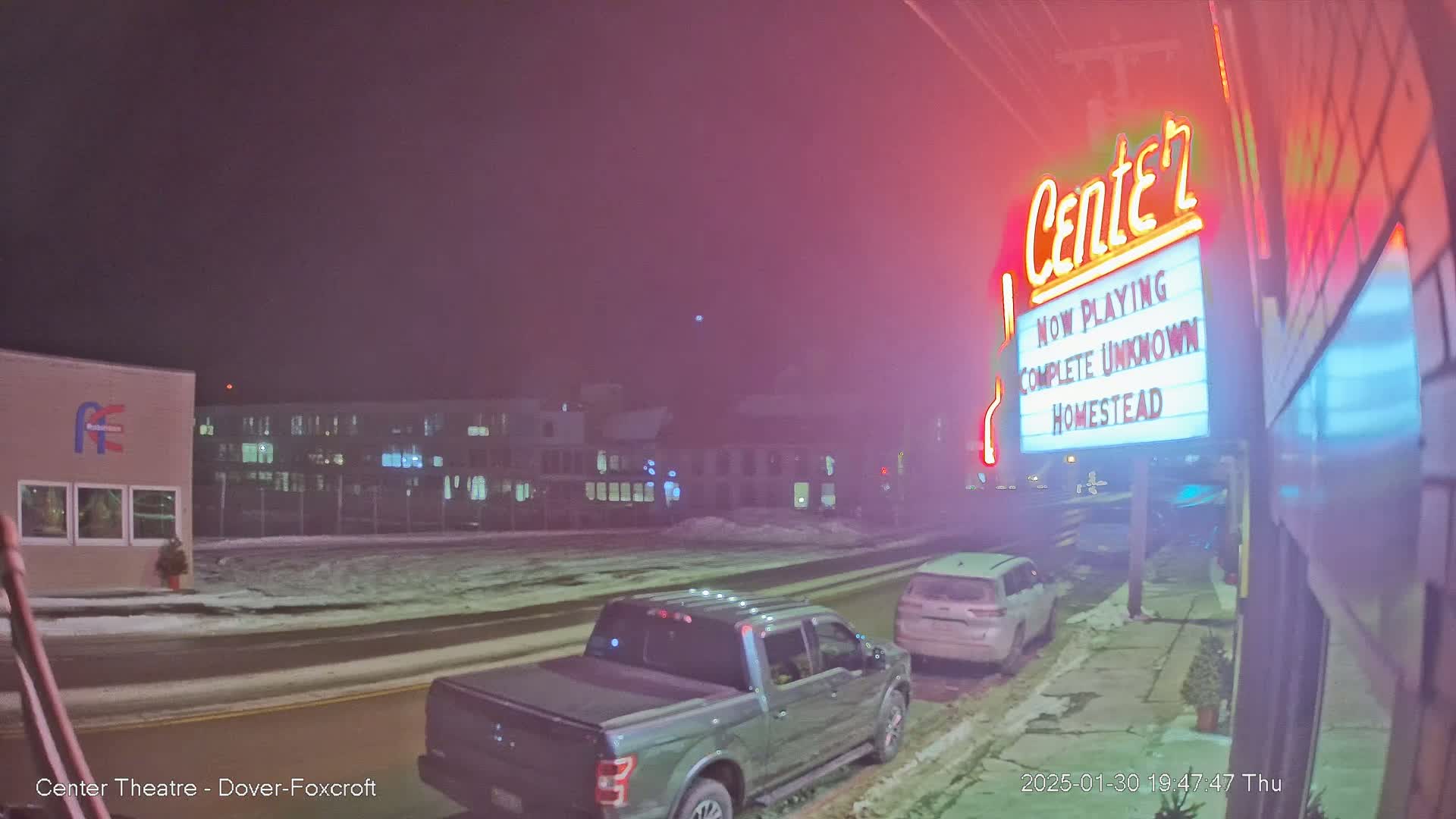 A snowy street at night shows a movie theater marquee advertising "Complete Unknown" and "Homestead," with a pickup truck and SUV parked nearby.