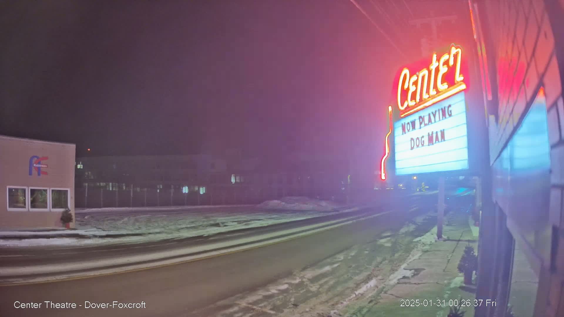 A snow-covered street at night leads to a movie theater marquee advertising "Dog Man," illuminated by neon lights in a foggy atmosphere.