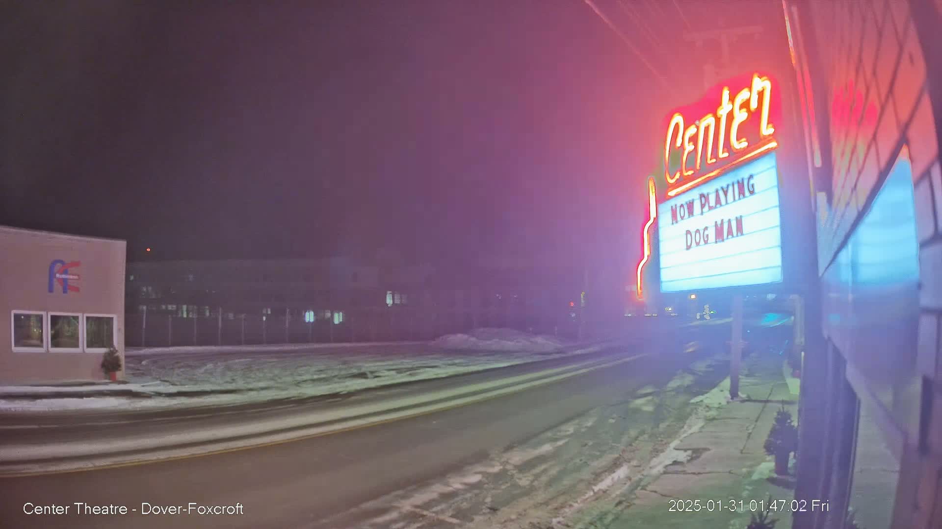 A snow-covered street at night, showing a movie theater marquee advertising "Dog Man," under a hazy, pink-tinged sky.