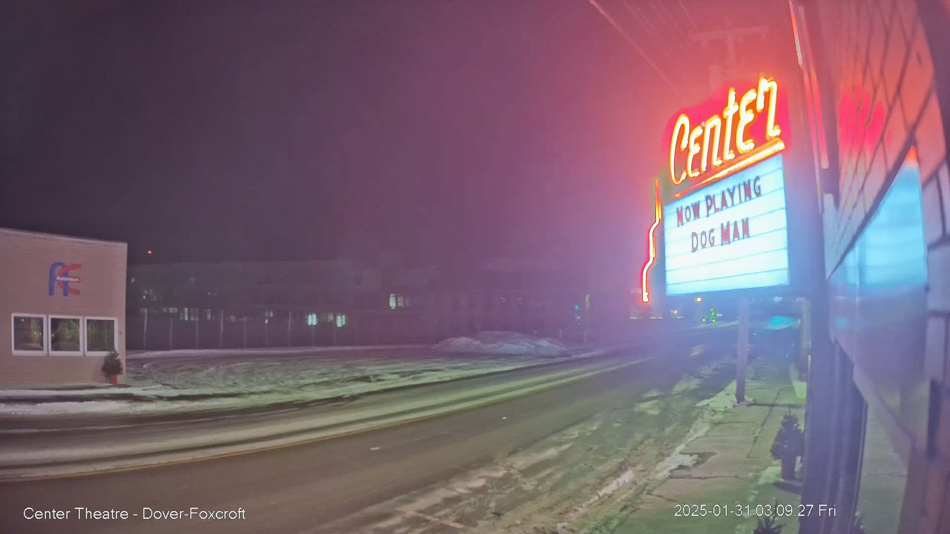 A snowy street at night is dimly lit, showing a movie theater marquee advertising "Dog Man" under hazy conditions.