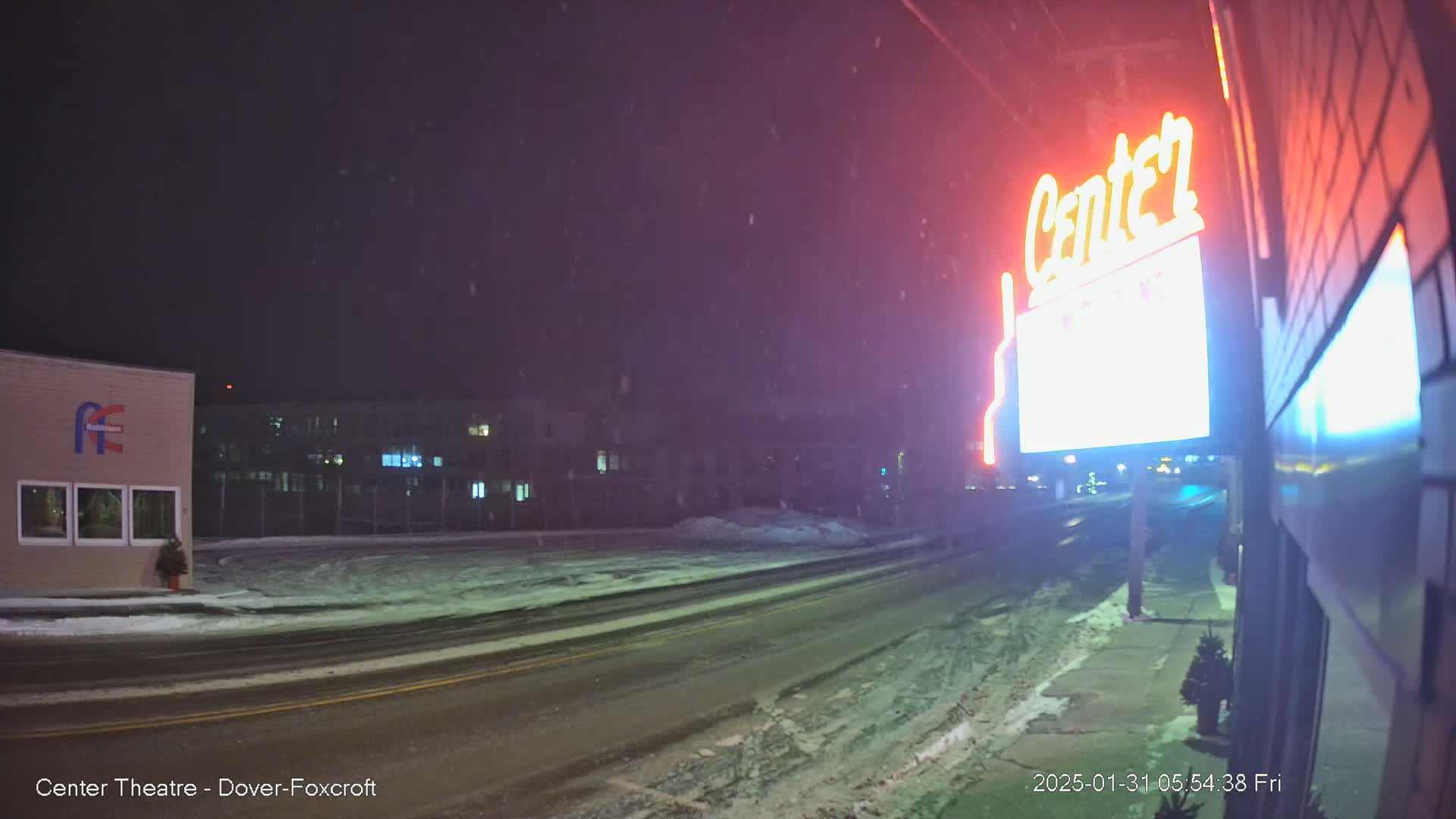 A snow-covered street at night, illuminated by a neon sign, with buildings and a small tree visible in the background under a lightly snowing sky.