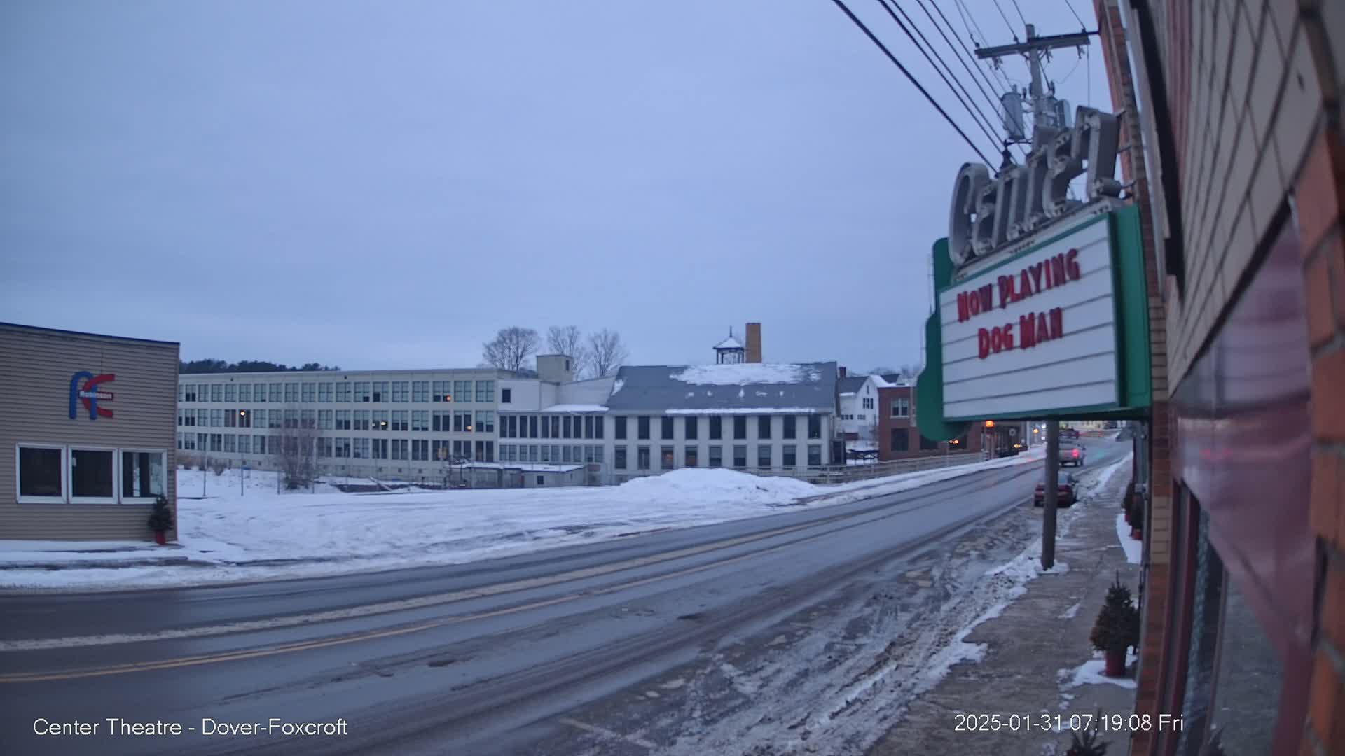A snow-covered street scene shows several buildings, including a large multi-story structure and a movie theater marquee, under a twilight sky.