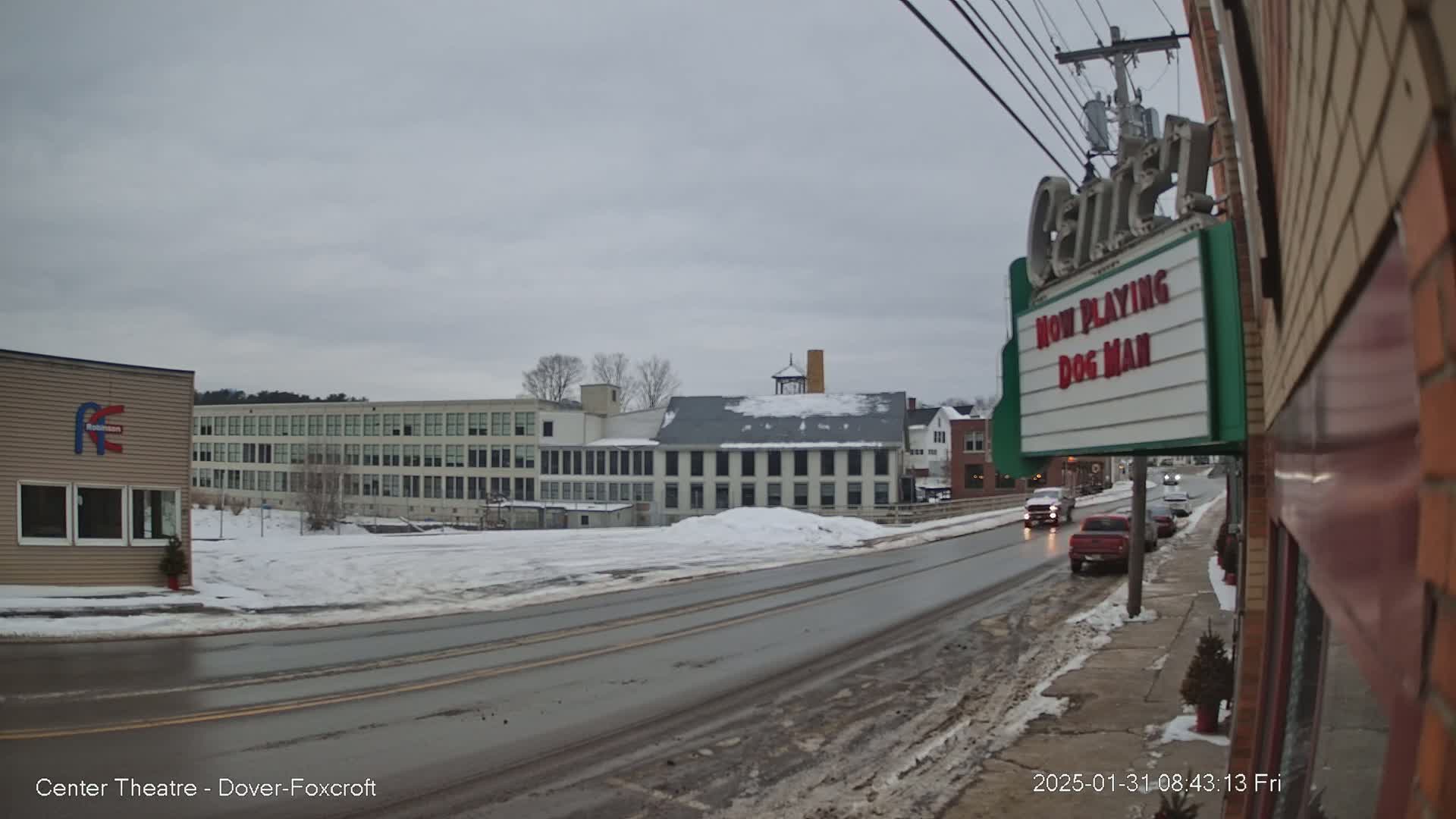 A snow-covered street scene shows a two-lane road with cars, several buildings including a large factory and a movie theater marquee, under an overcast sky.