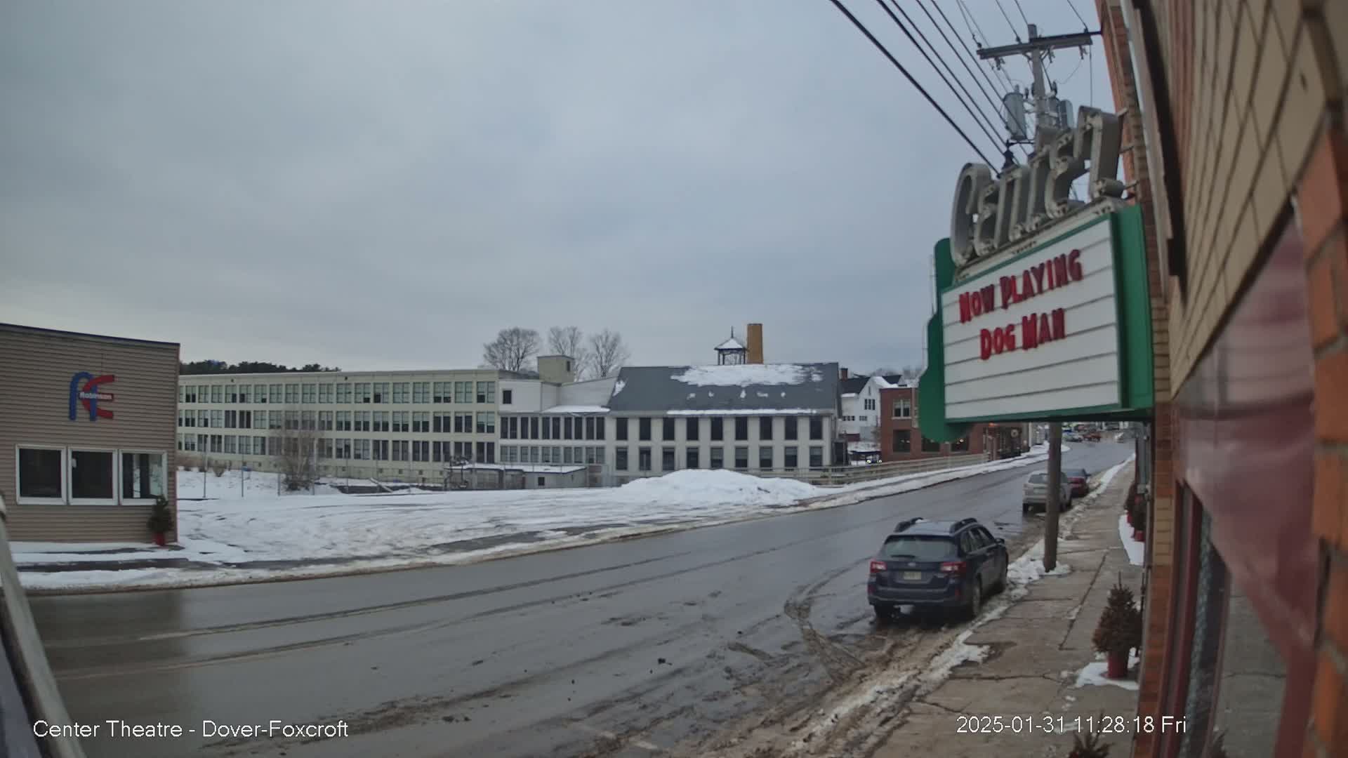 A snow-covered street scene shows several buildings, including a large multi-story building and a movie theater marquee displaying "Now Playing Dog Man," under an overcast sky.