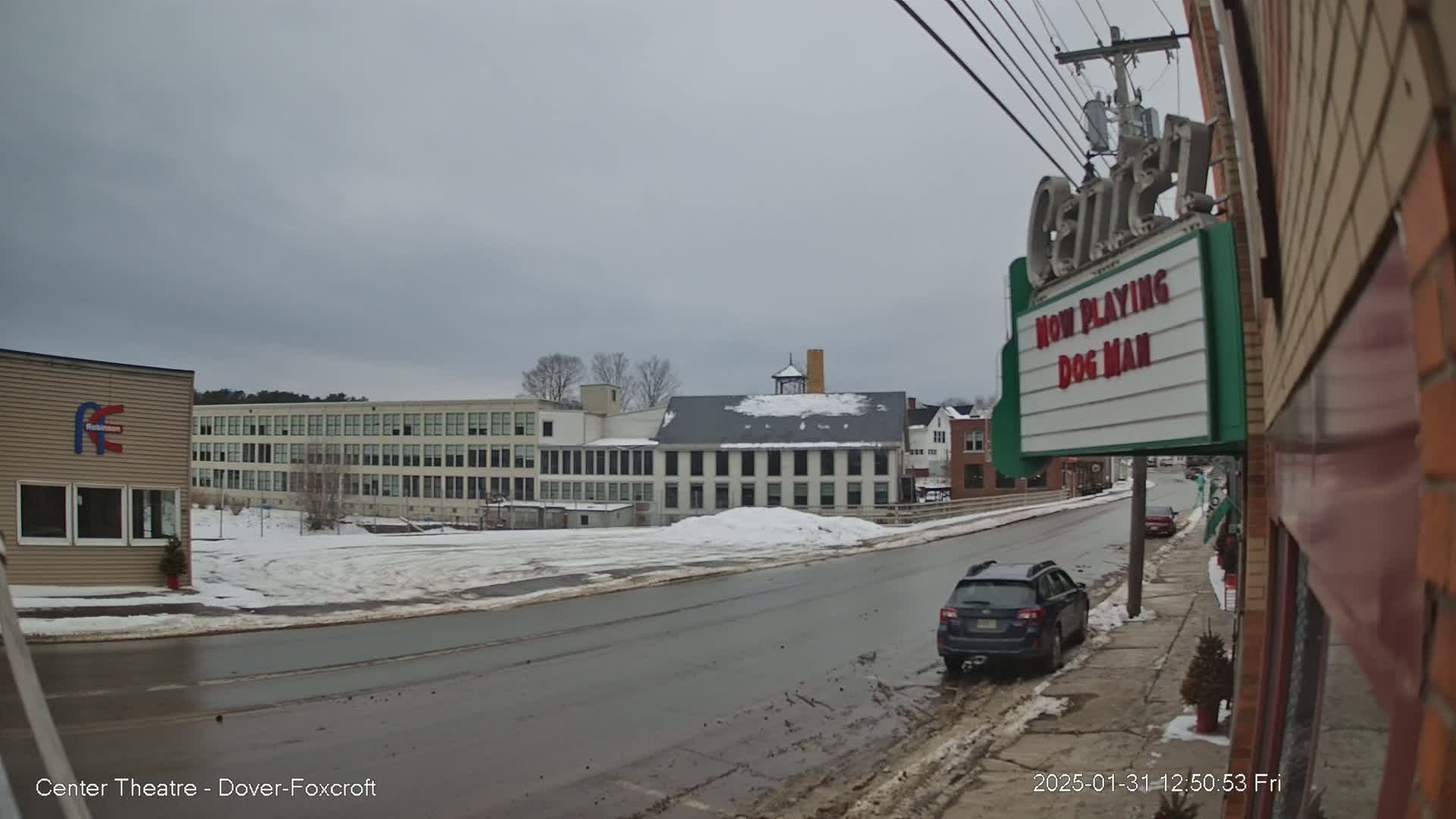 A snow-covered street scene shows several buildings, including a large factory-like structure and a movie theater marquee advertising "Dog Man," under a cloudy sky.