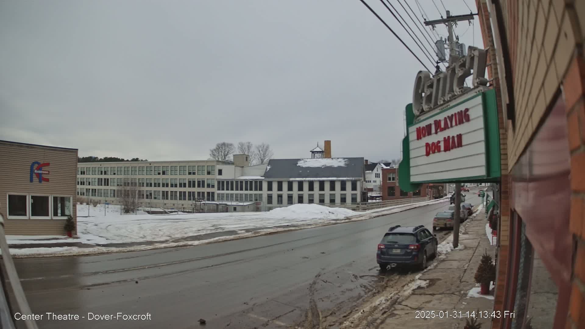 A snow-covered street scene shows several buildings, including a large factory and a movie theater marquee advertising "Dog Man," under an overcast sky.