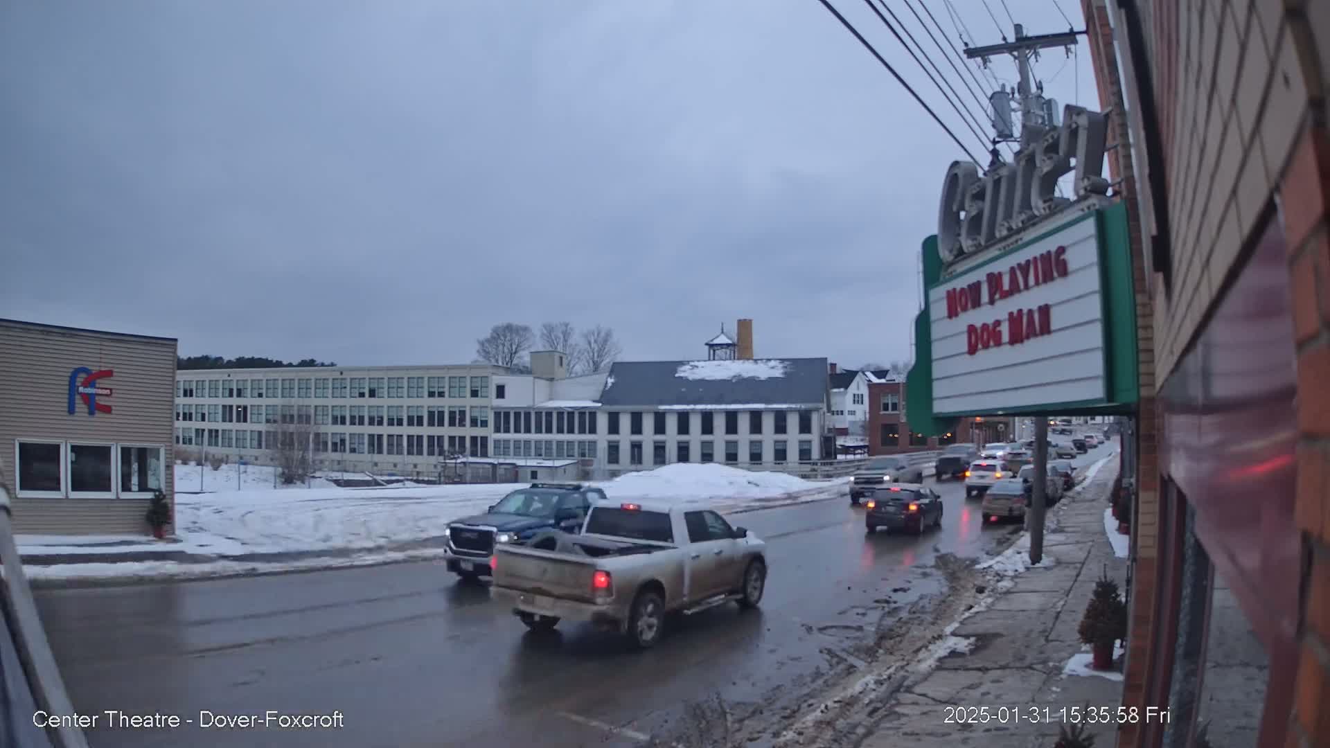 A street scene on an overcast day shows several vehicles driving on a wet, snow-covered road past a multi-story building and a movie theater marquee.