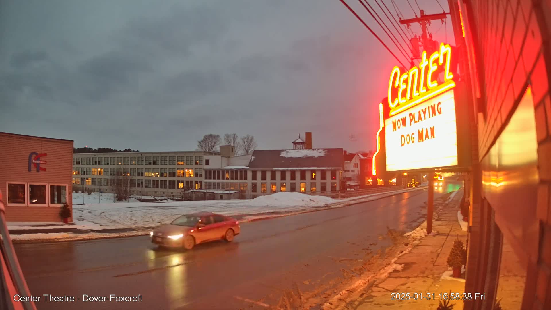 A red car drives past a movie theater marquee displaying "Dog Man" under a dark, overcast sky on a wet street with some snow on the ground.