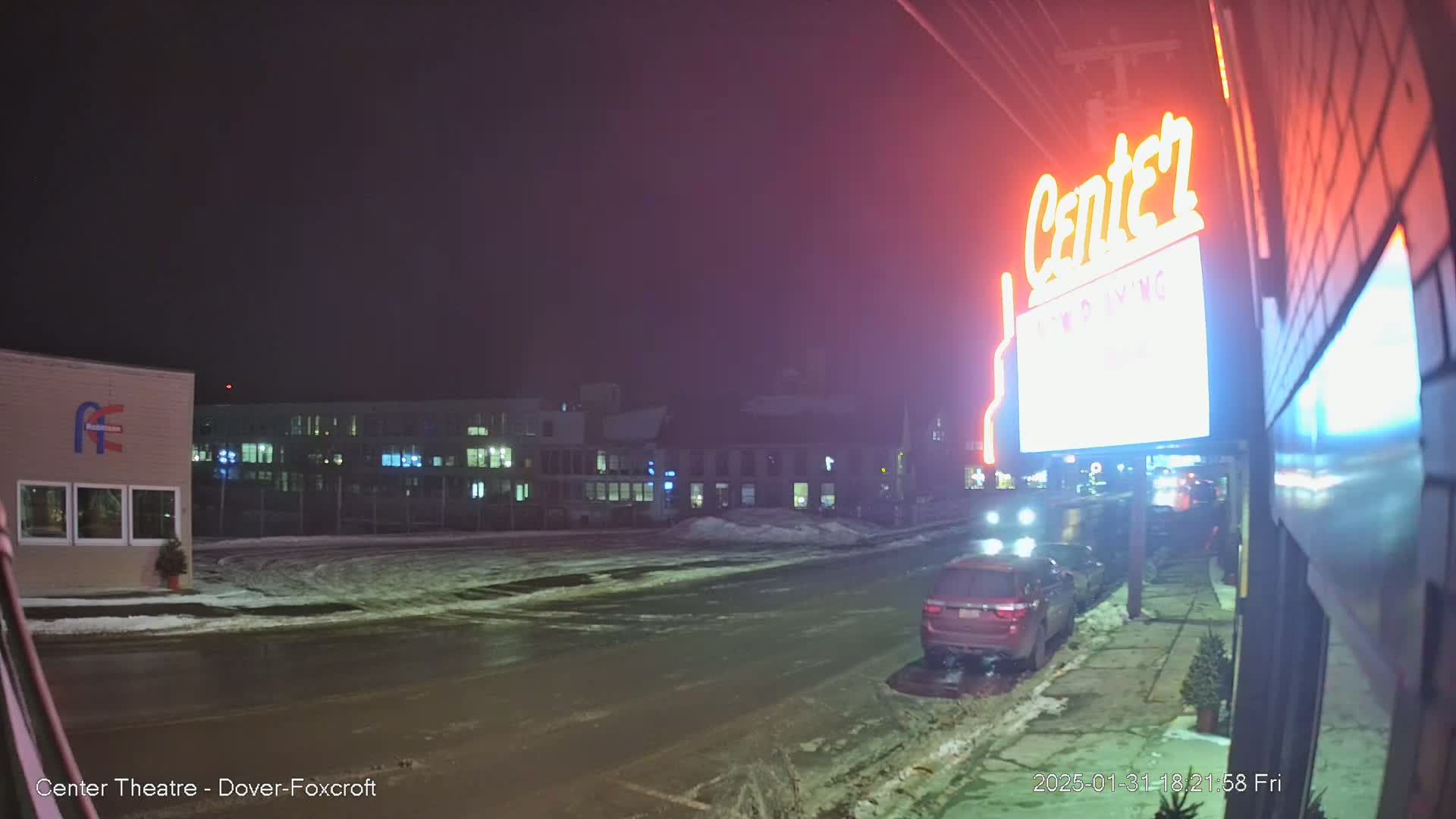 A snow-covered street at night shows a red SUV parked near a building with a lit "Center" sign, other buildings in the background, and some cars in the distance.