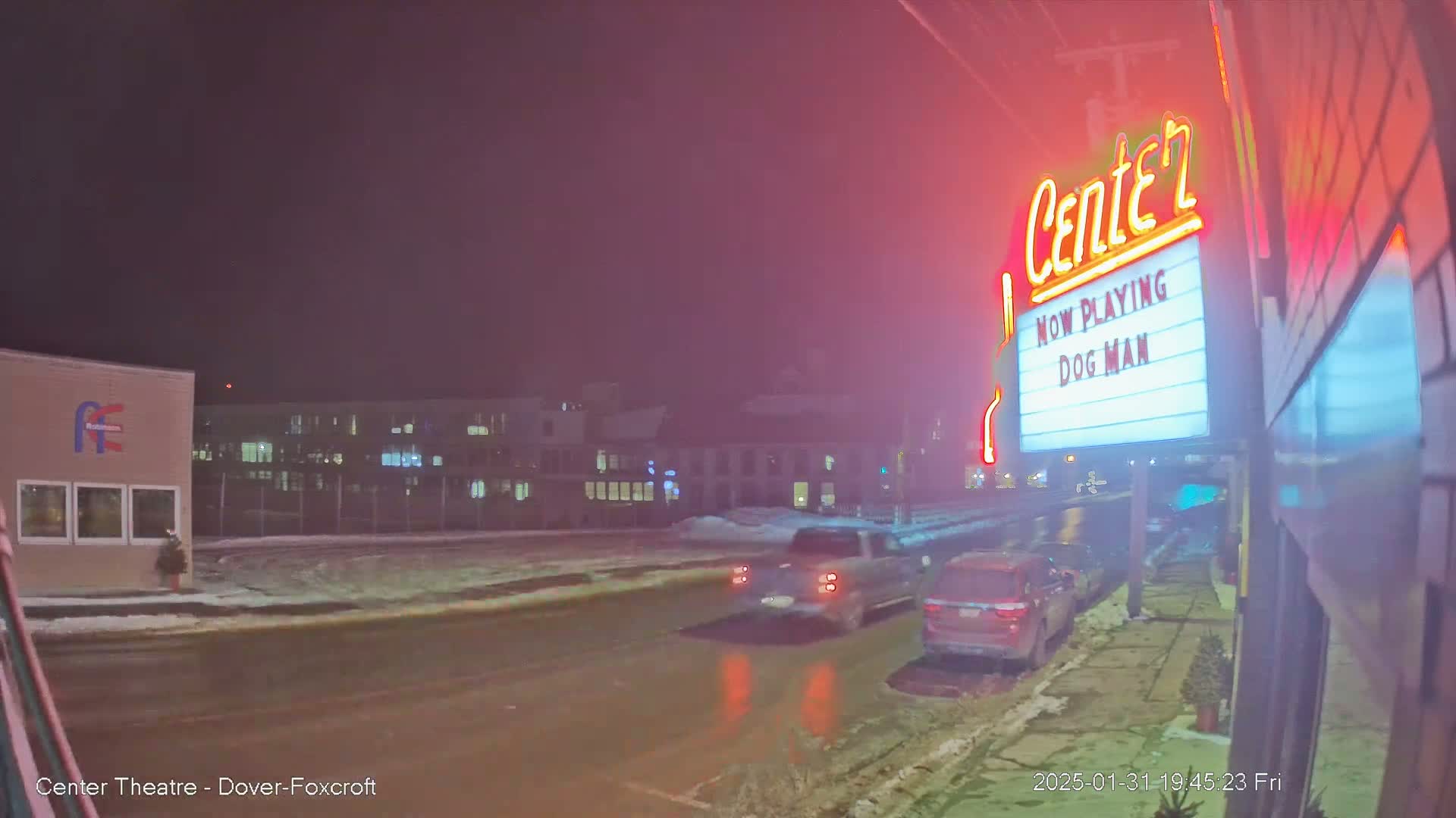 A snowy, nighttime street scene shows a movie theater marquee advertising "Dog Man," with several vehicles parked nearby.