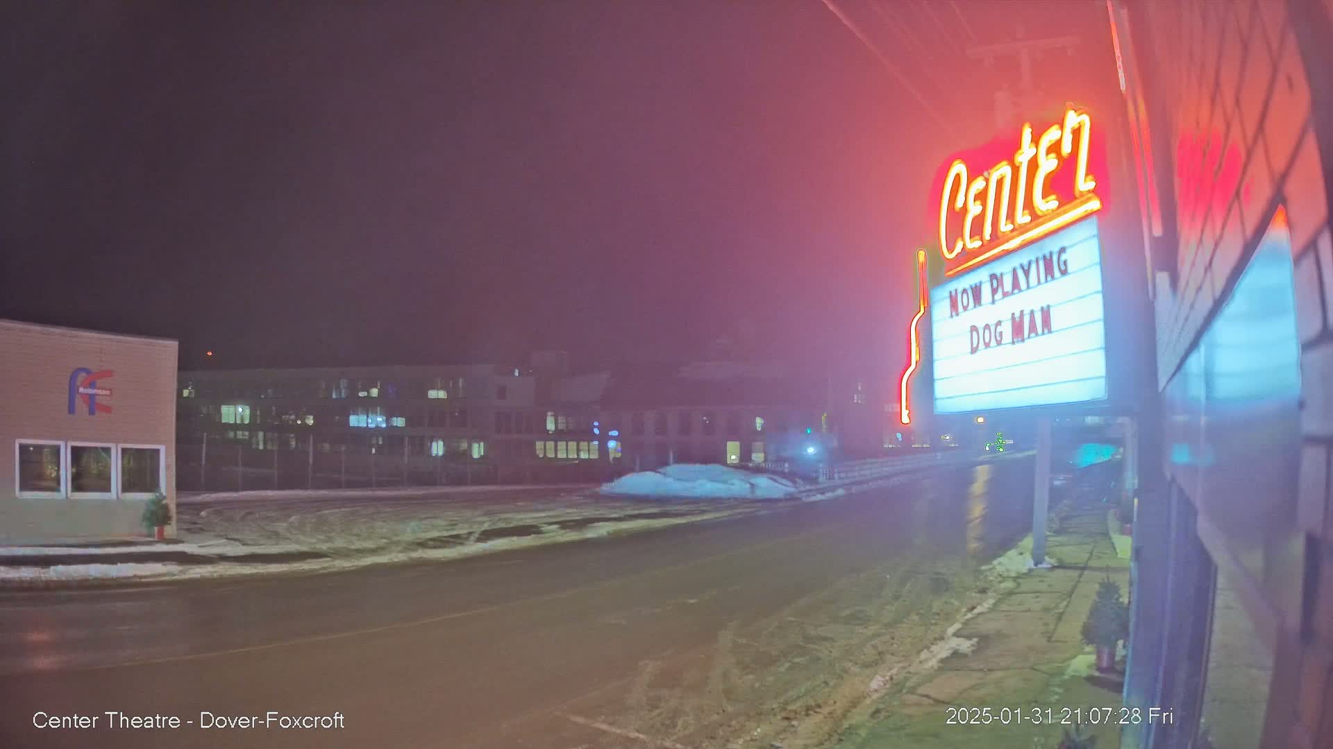 A nighttime, snowy street scene shows a movie theater marquee advertising "Dog Man," with buildings and a partly snow-covered road in the background under a hazy, dimly lit sky.