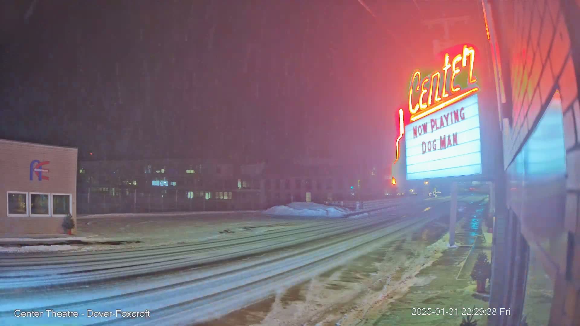 A snow-covered road at night, showing a movie theater marquee advertising "Dog Man," with buildings and some snowdrifts visible in the background.