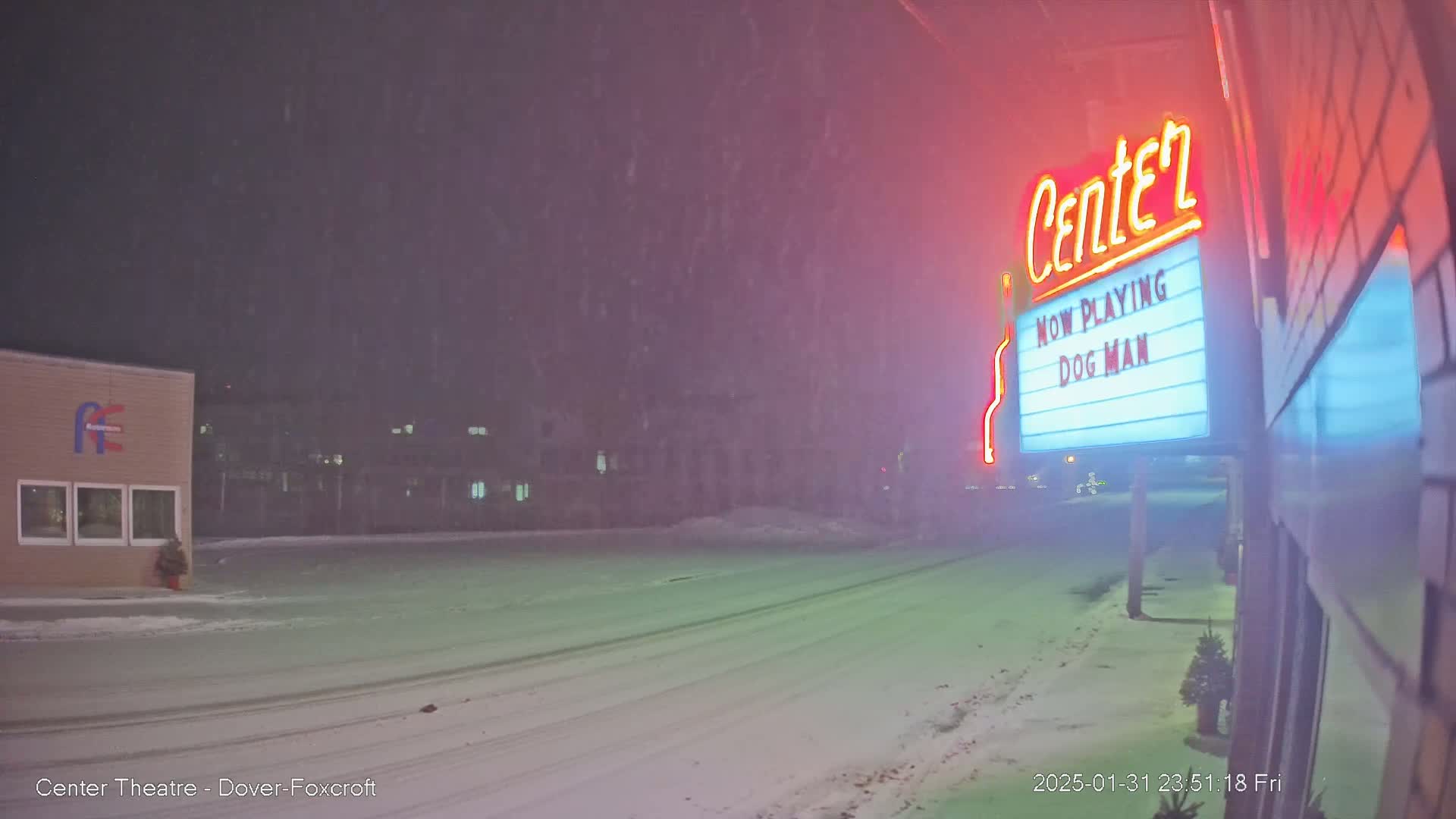 A snow-covered street at night shows a movie theater marquee advertising "Dog Man" under a falling snow.