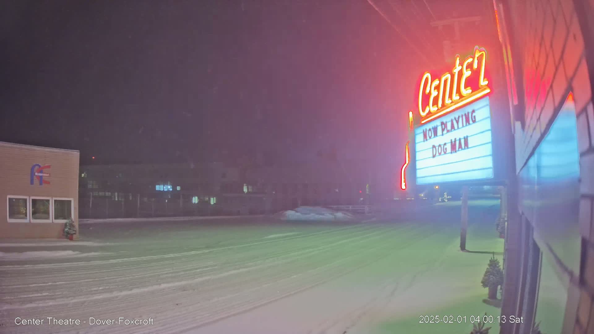 A snow-covered parking lot at night with a movie theater marquee showing "Dog Man" illuminated by neon lights.
