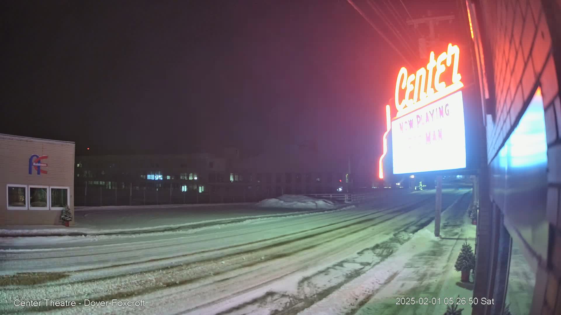 A snow-covered street at night is seen from a street-level perspective, with a brightly lit theater marquee in the foreground and buildings in the background.