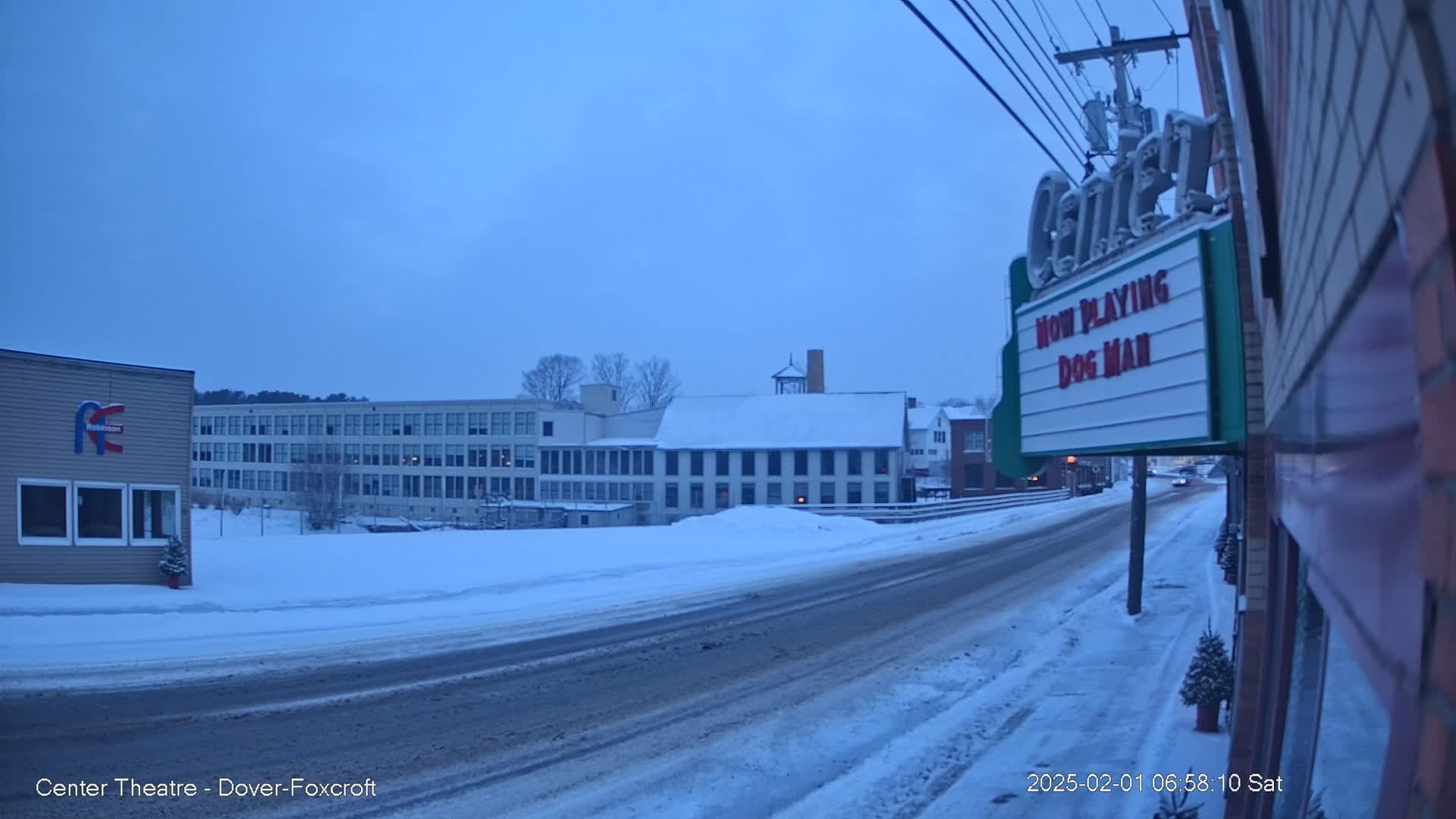 A snow-covered street scene shows several buildings under a twilight sky, with a road curving toward a movie theater marquee.