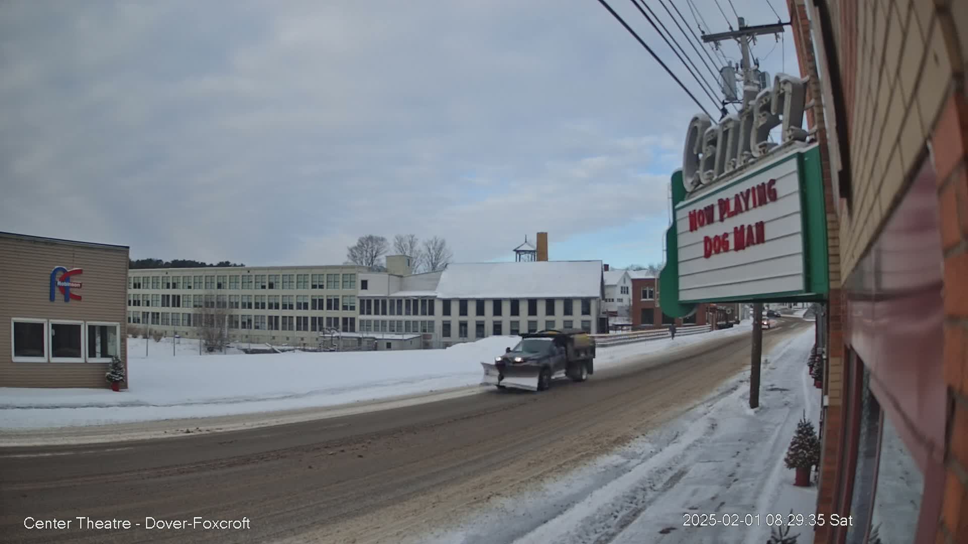 A snow-covered street scene shows a snowplow driving past buildings, including a large factory and a movie theater marquee.