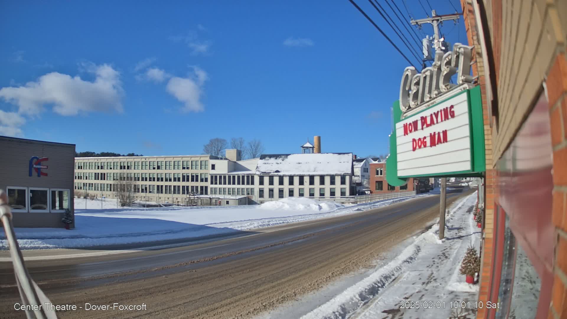 A snow-covered street scene shows several buildings, including a large factory and a movie theater marquee advertising "Dog Man," under a mostly sunny sky.