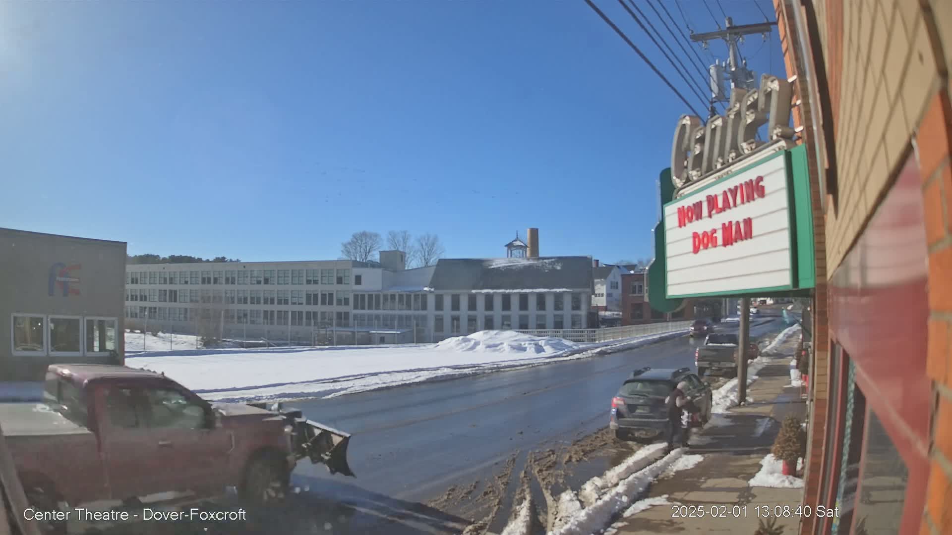 A sunny winter day reveals a street scene with snow-covered ground, a partially plowed road, vehicles parked along the curb, and buildings including a school and a movie theater showing "Dog Man".