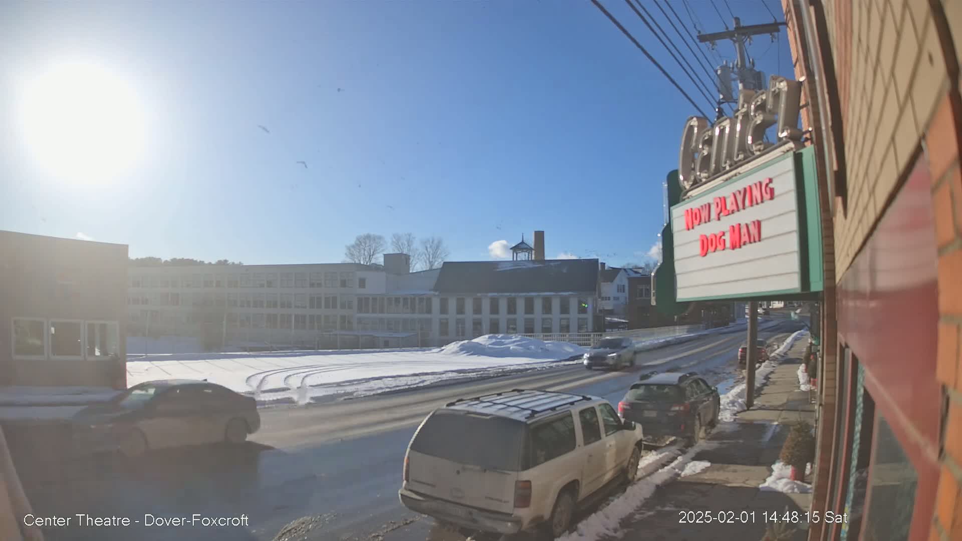 A sunny winter day scene shows a snow-covered street with cars, a large building, and a movie theater marquee.