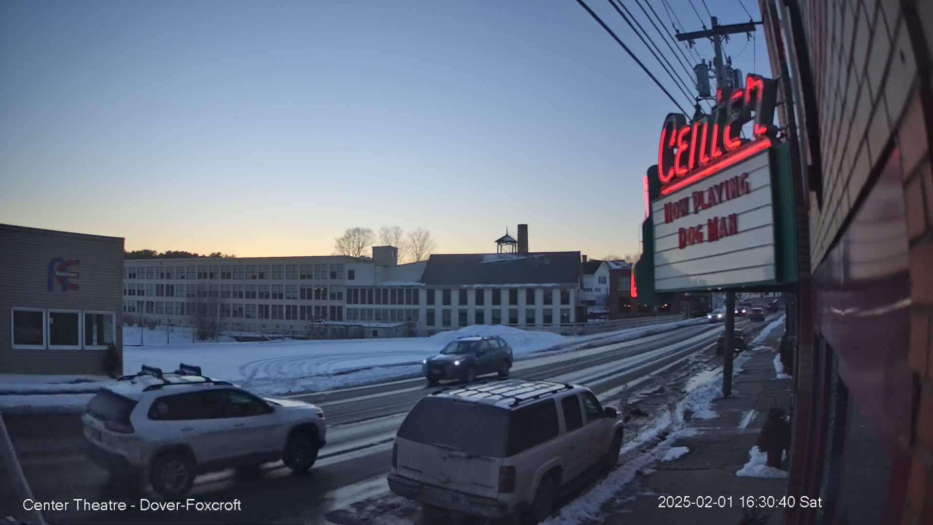 A snow-covered street scene at dusk shows several vehicles traveling on a road past buildings, one of which is a movie theater marquee advertising "Dog Man."