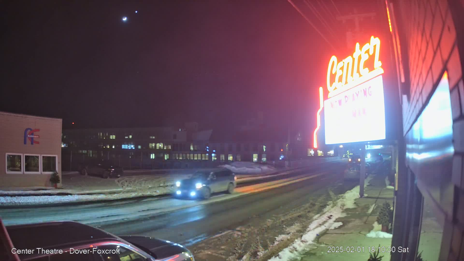 A snowy nighttime street scene shows a lit-up theater marquee and several vehicles driving past under a dark sky with a bright moon and stars.