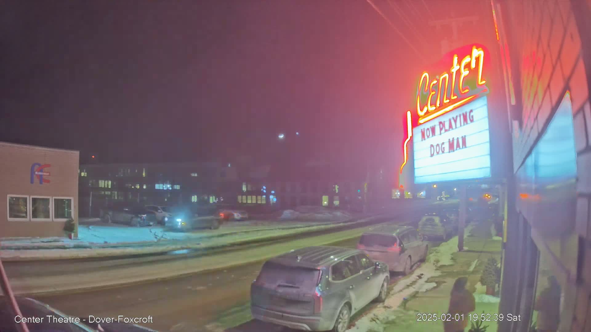 A snowy street at night shows several parked cars in front of a movie theater marquee advertising "Dog Man."