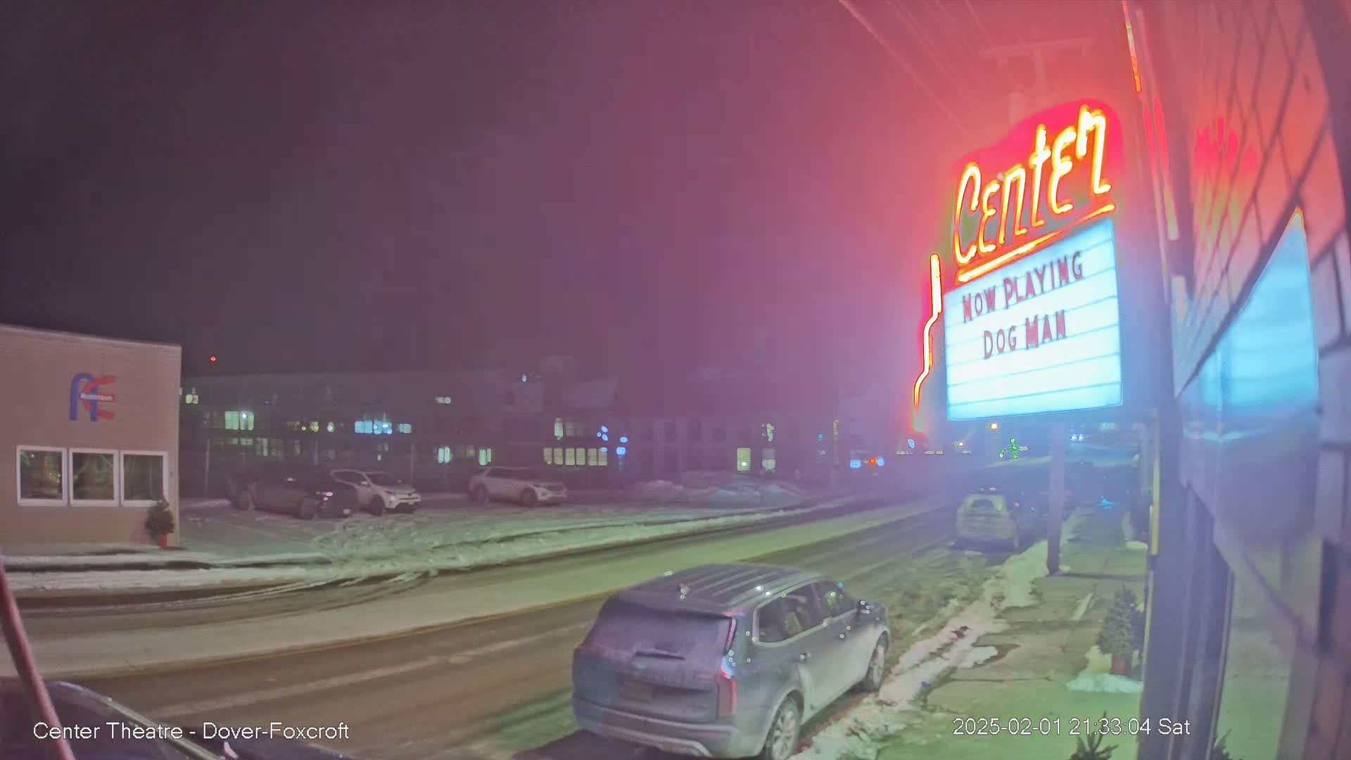 A snow-covered street at night shows a movie theater marquee advertising "Dog Man," with cars parked along the side of the road and a vehicle driving away.