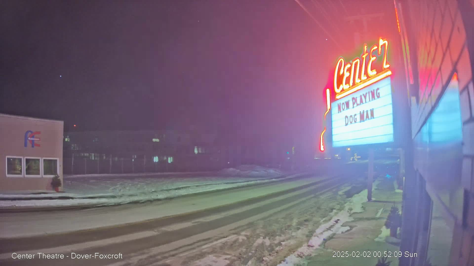 A snow-covered road at night leads to a movie theater marquee advertising "Dog Man," under hazy, pinkish-red nighttime conditions.