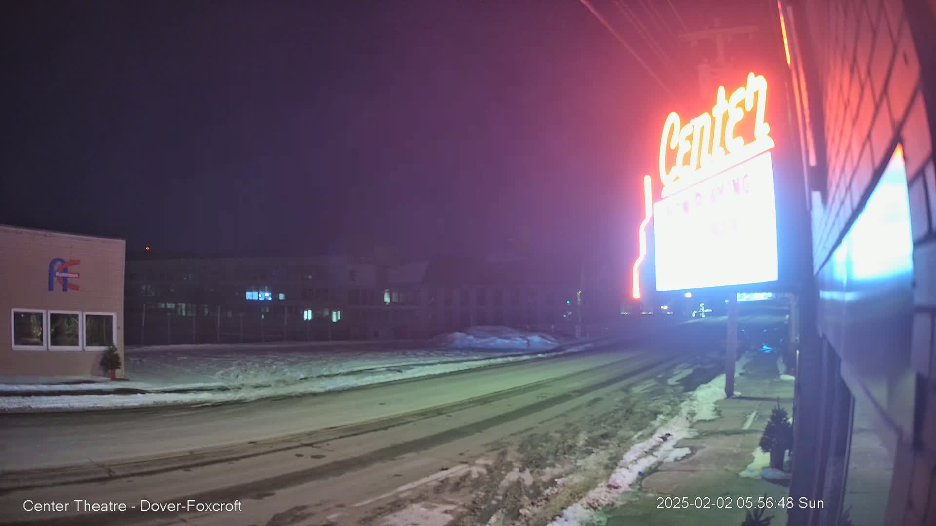 A nighttime street scene shows a snow-covered road in front of a building with a neon "Center" sign, and other buildings are visible in the background under a dark sky.