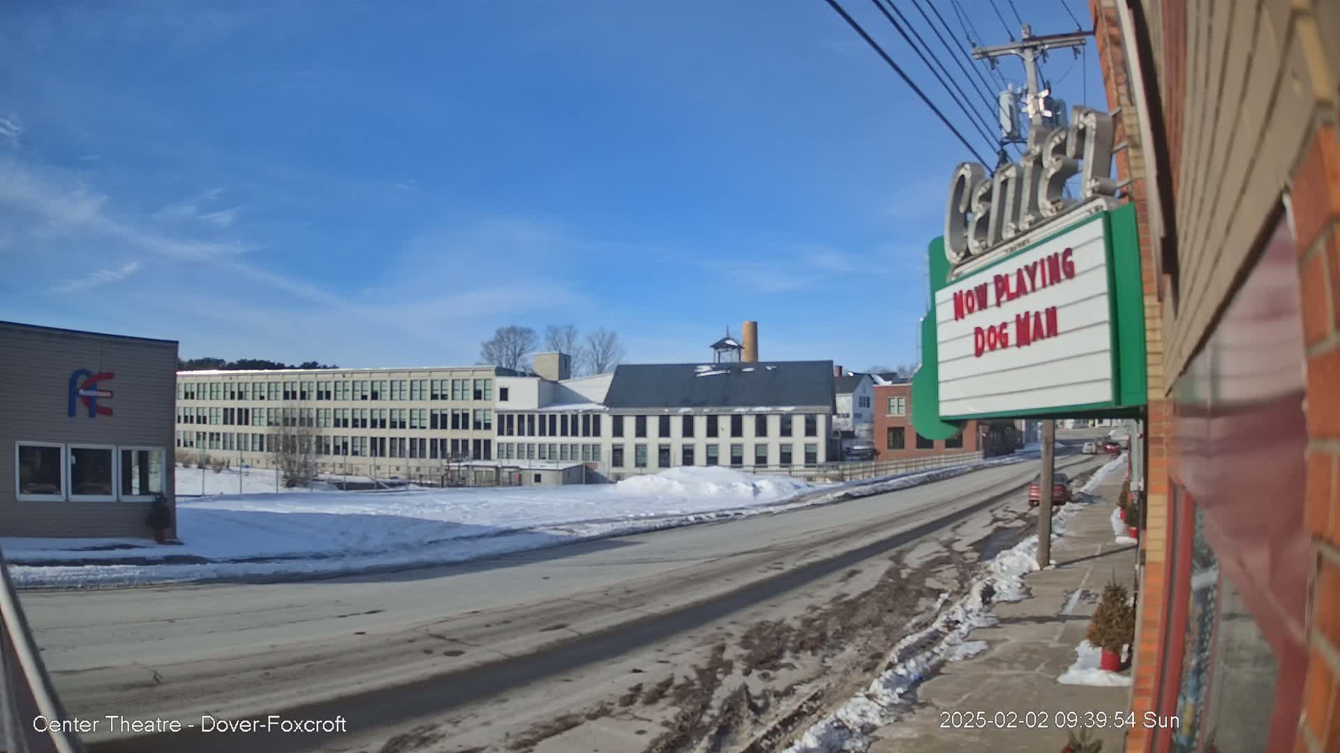 A sunny winter day reveals a street scene with snow-covered ground, multi-story buildings, and a movie theater marquee.