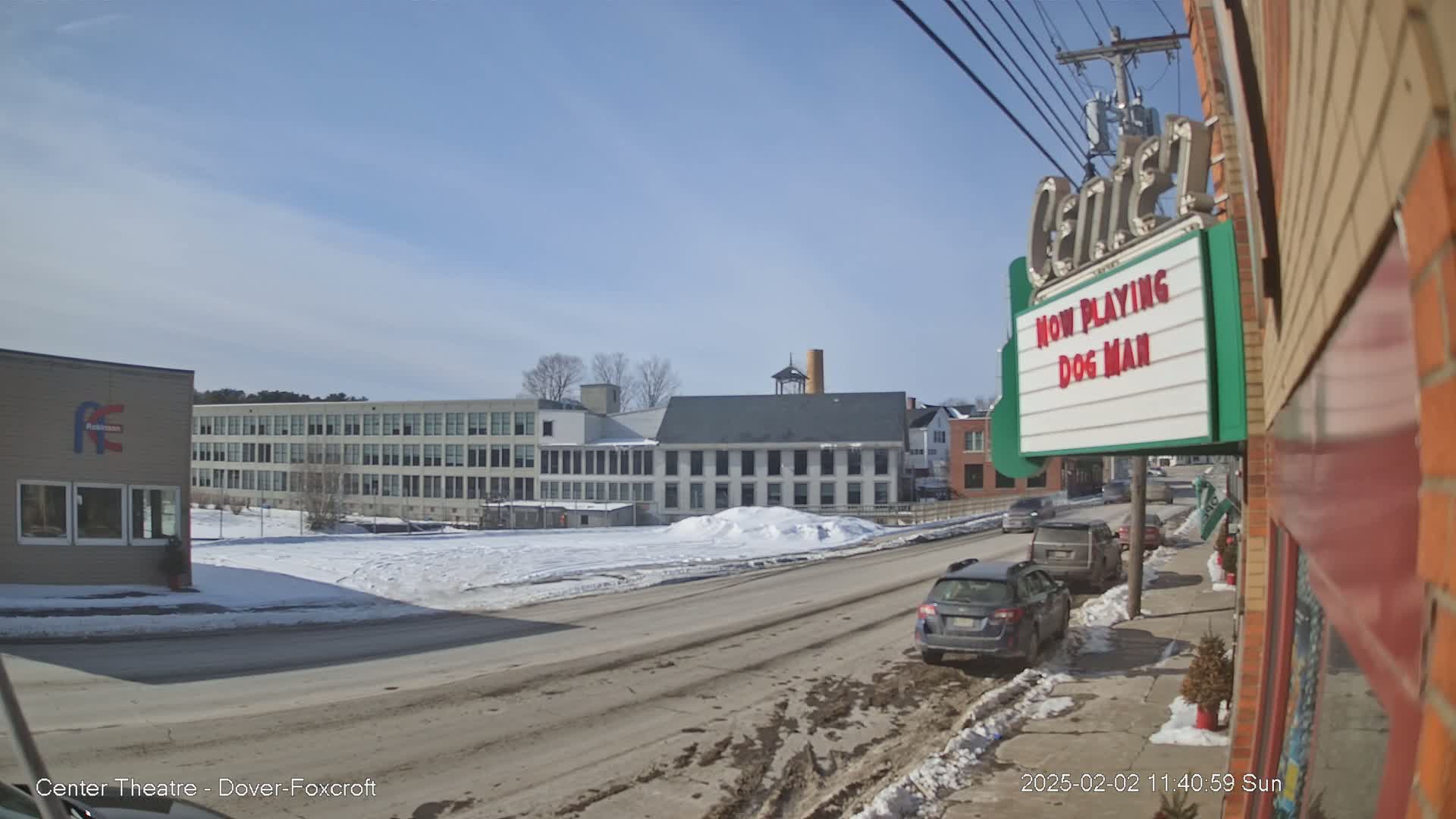 A sunny winter day reveals a street scene with parked cars, a multi-story building in the background, and a movie theater marquee advertising "Dog Man."